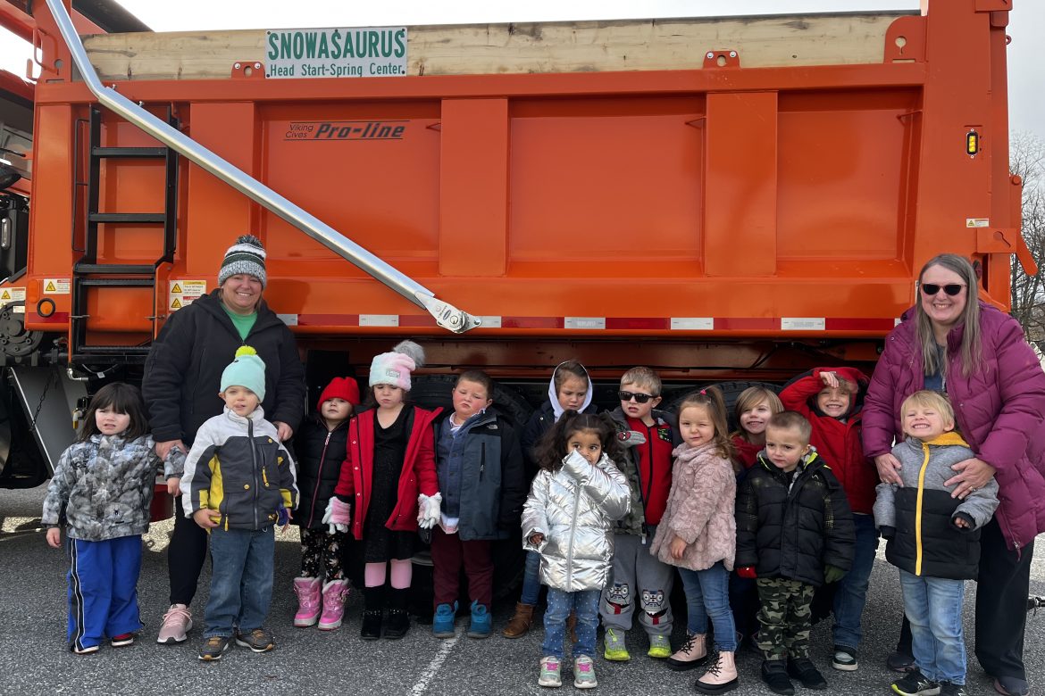 A group of school-aged children dressed in winter clothes standing with their teachers in front of a parked plow truck.