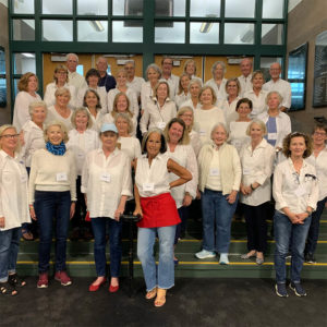A large group of mostly women pose on stairs for picture. They are all wearing white shirts and blue jeans with name tags hanging around their necks.