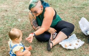A woman with blue hair sits on a grass feild and hands a baby a small peice of bread from a sandwich she is eating. 