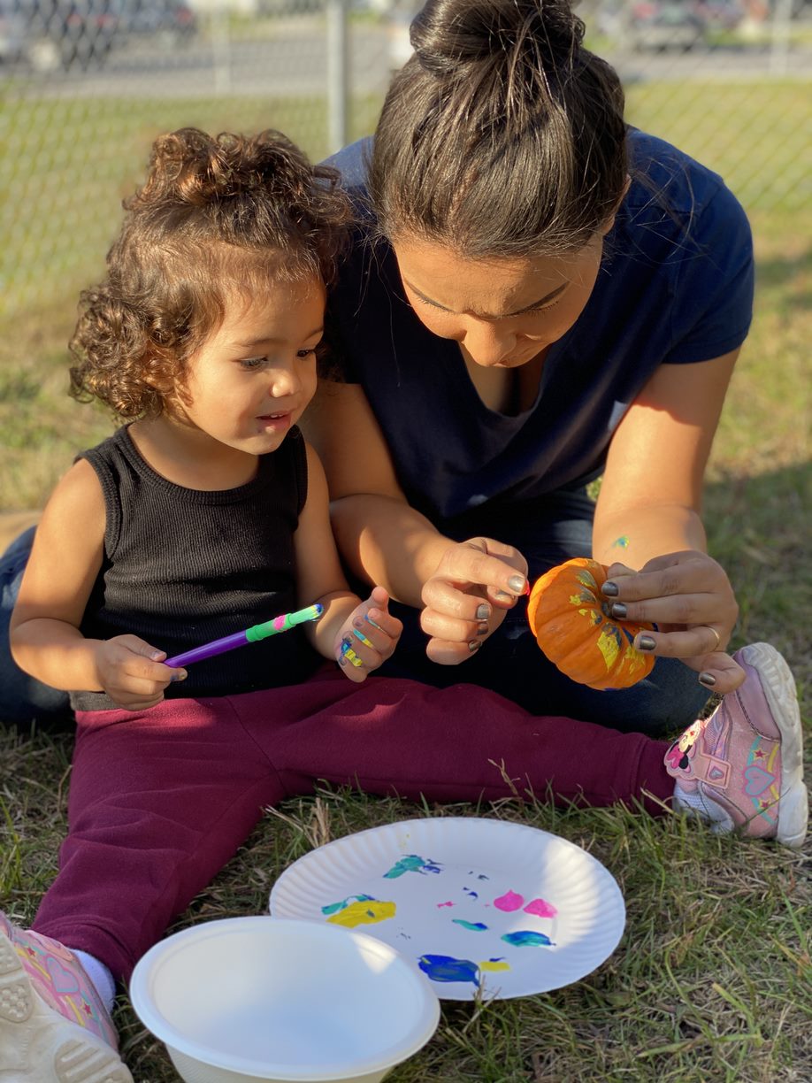 A mother helps a young girl paint a pumpkin outside while sitting in the grass. 
