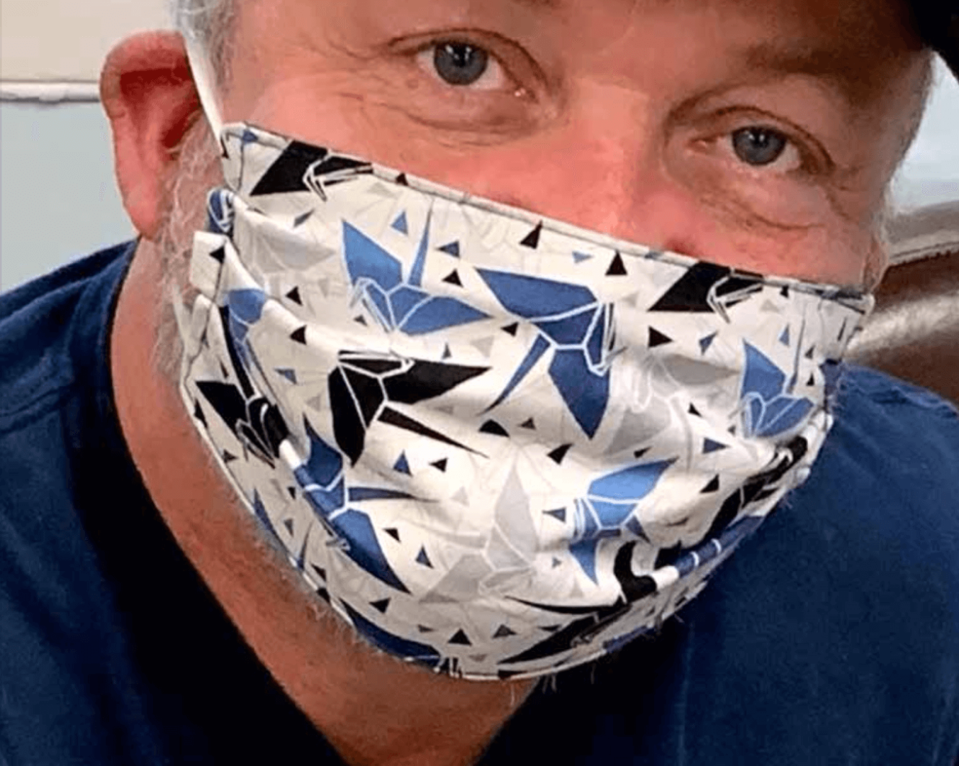 A close up image a man's face wearing a blue, white, and light blue mask. You can tell he's smiling behind the mask.