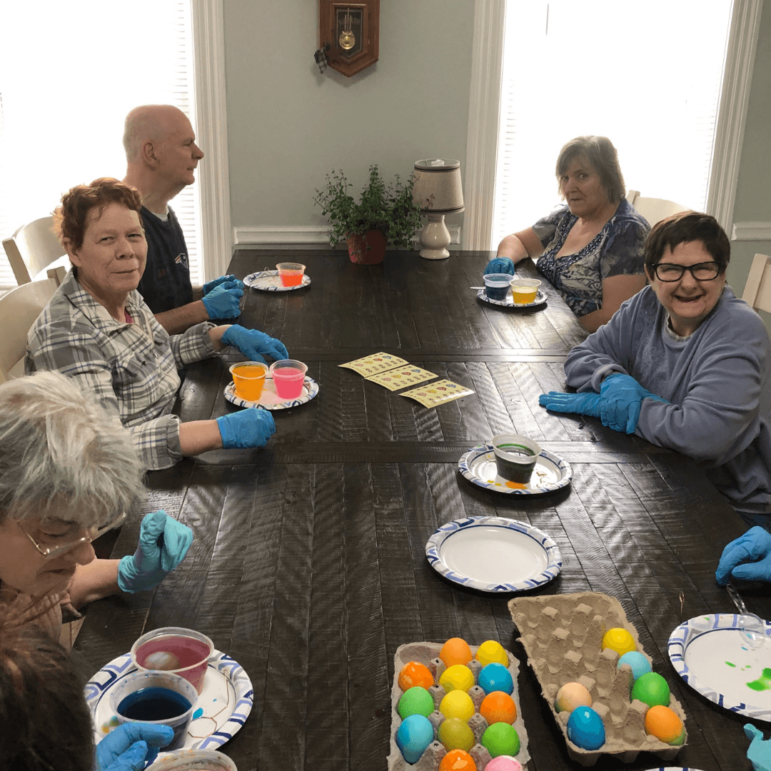 A group of adults sit around a dining room table decorating colorful Easter eggs.