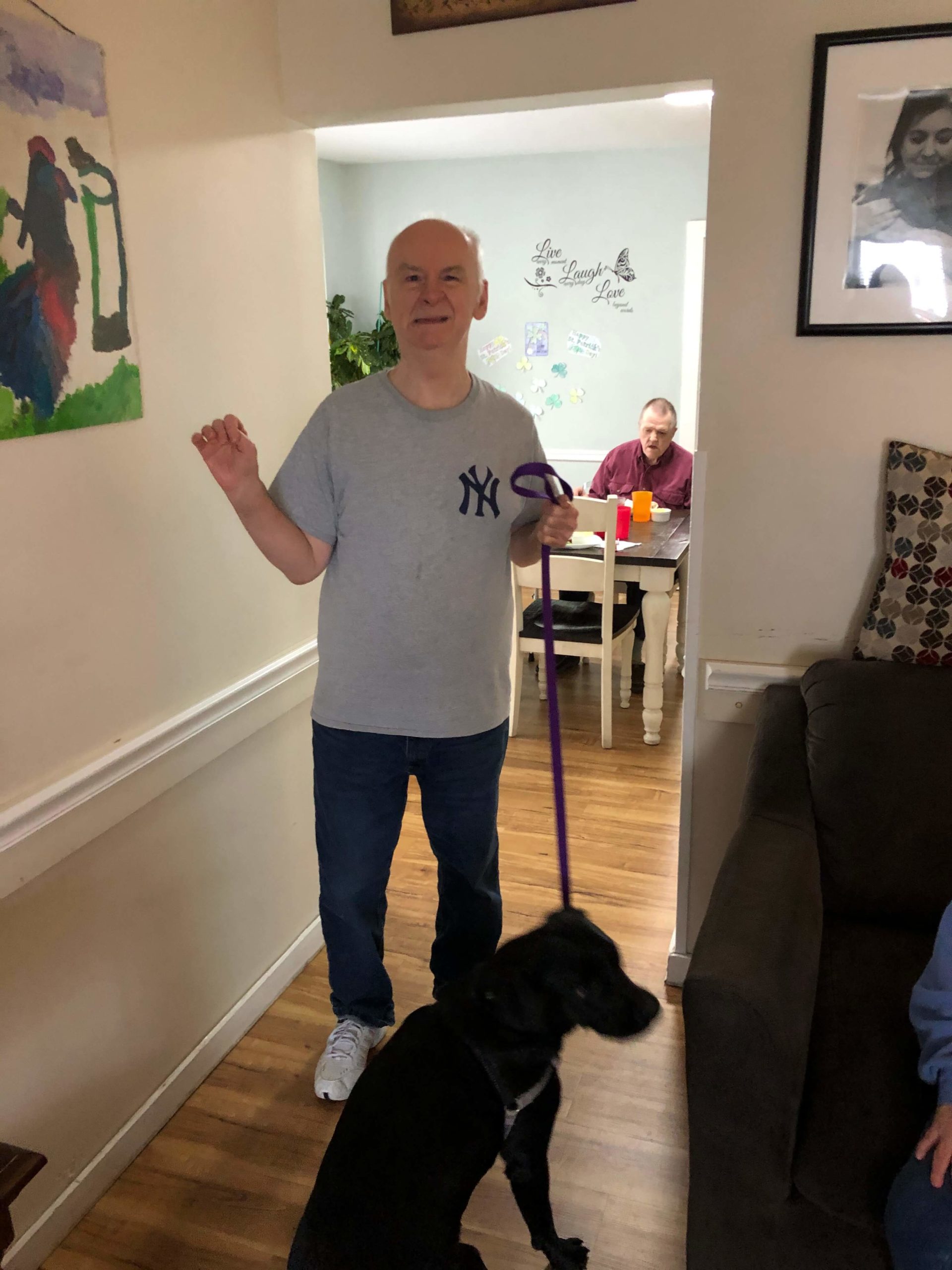 A man in a gray yankees t shirt hold the end of leash with a black dog attached to the other end. They are inside what appears to be a living room.