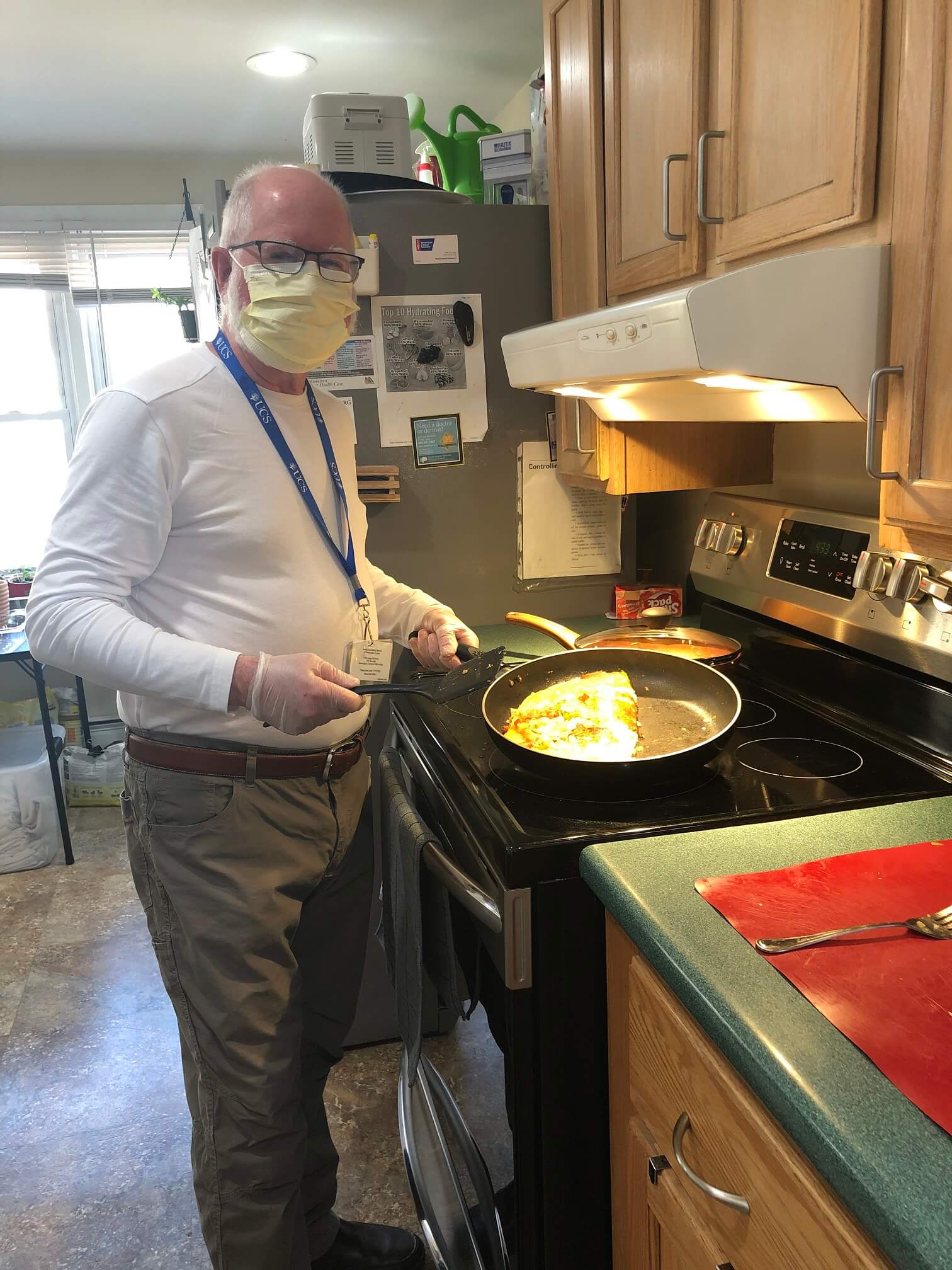 A man wearing a blue lanyard makes on omelet on a black stovetop. He is wearing face mask.