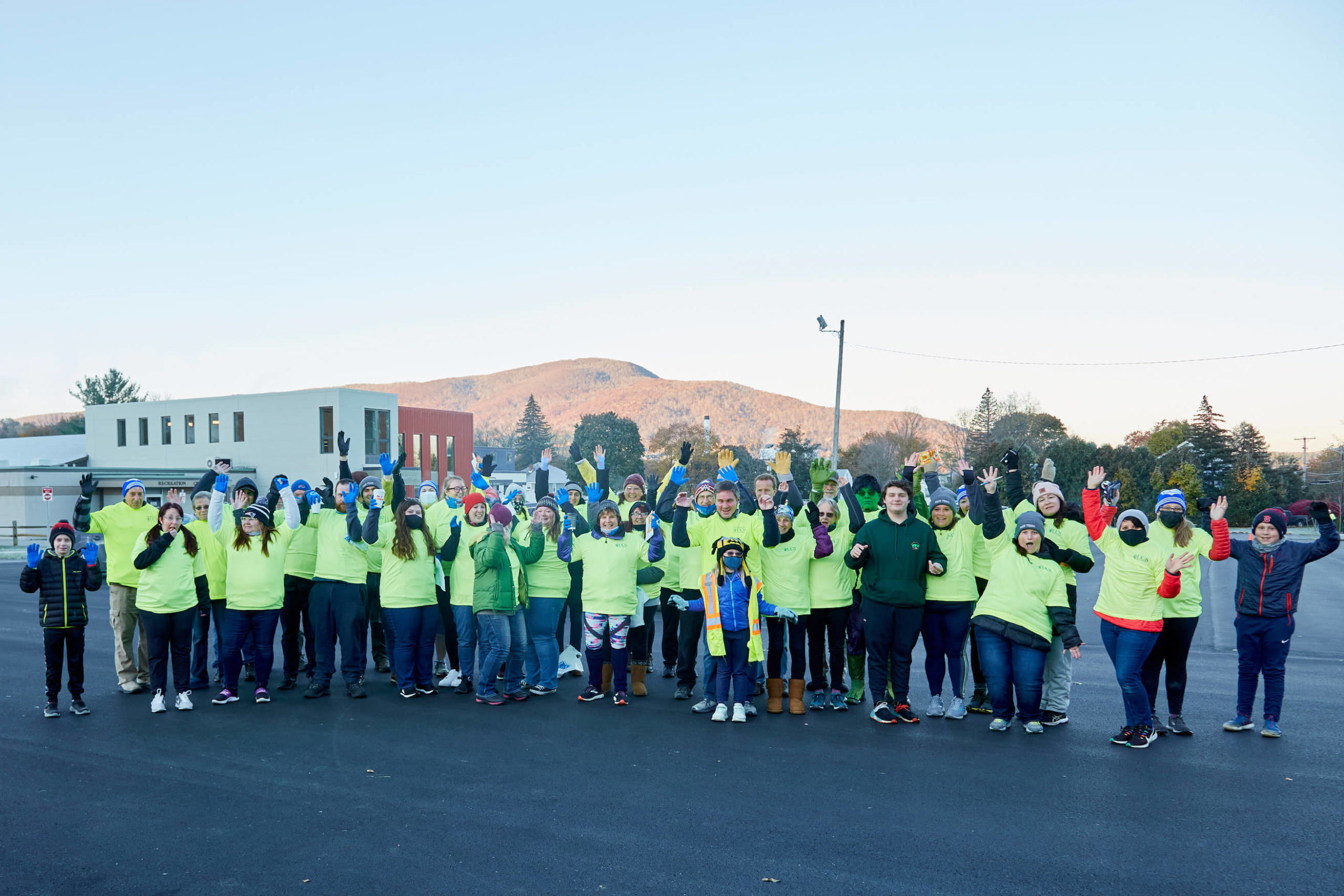 A large group of 5K volunteers wearing bright green shirts waves at the camera with Mt. Anthony and the UCS Spring Center in the background