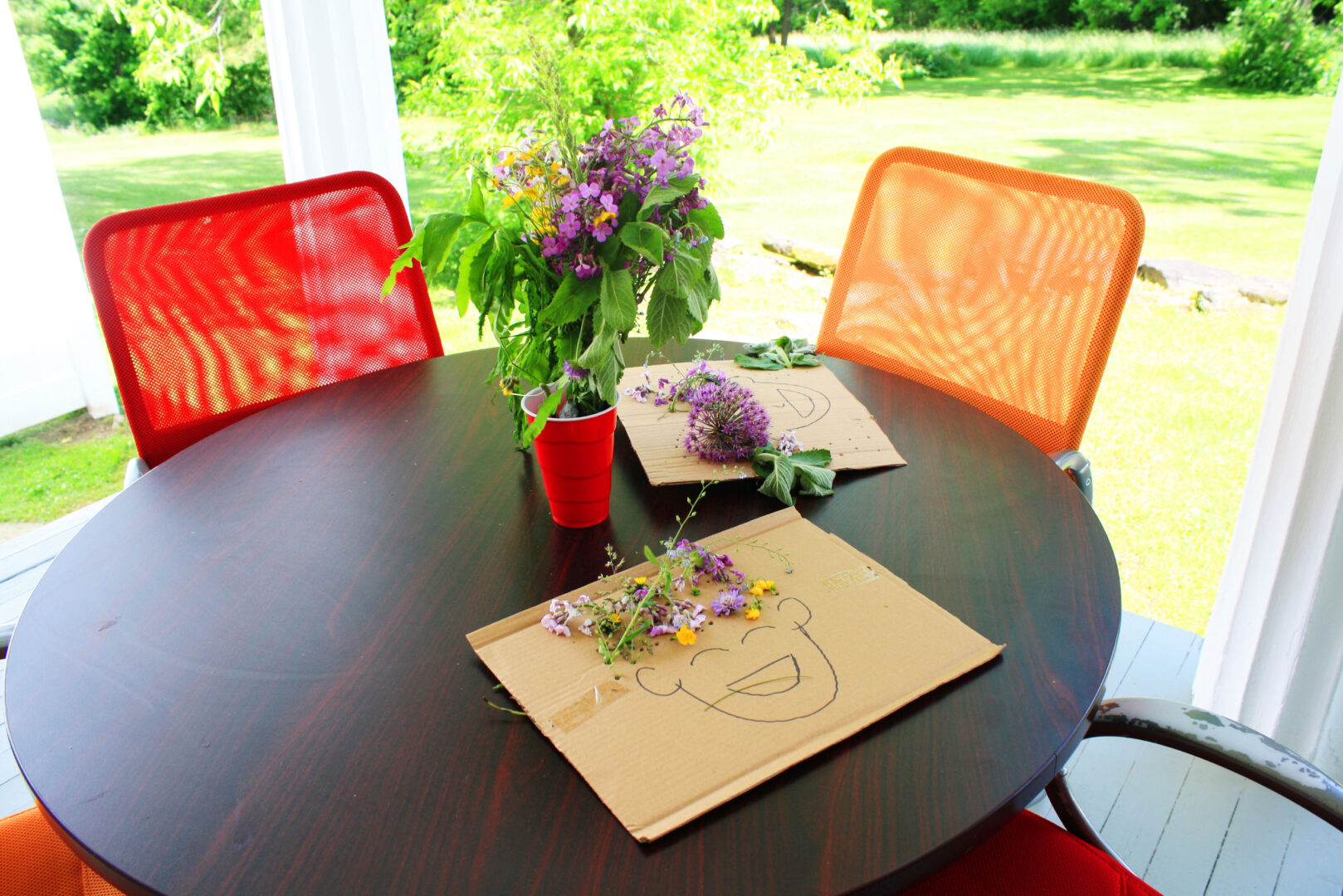 A round table outside holds cardboard artwork with flowers as hair on a person's face. Purple flowers in a red plastic cup on the table.