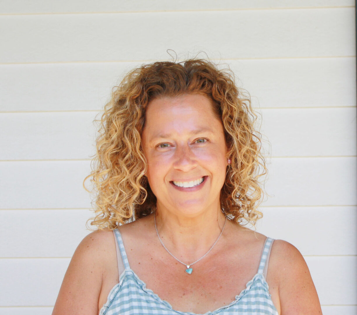 headshot of a woman with curly blonde hair wearing a spaghetti strap blue and white checkered dress smiles against a white clapboard background