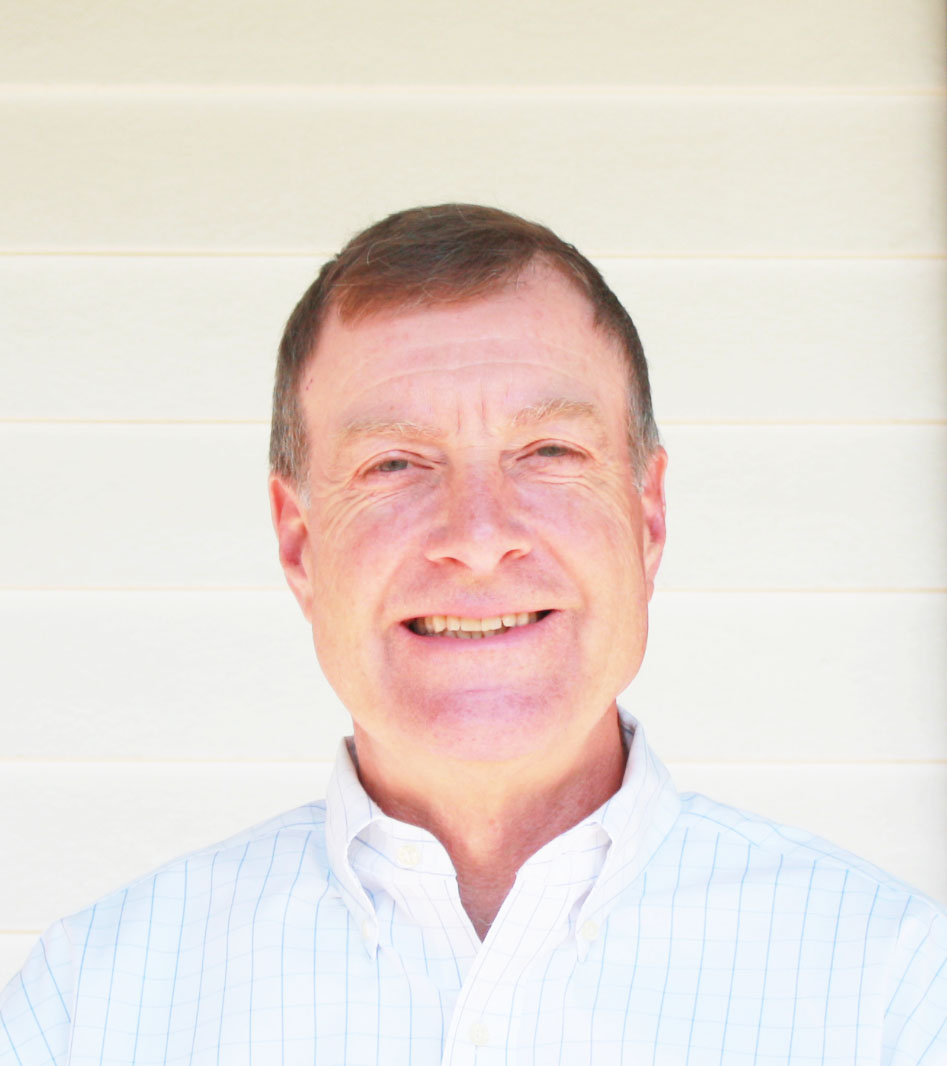 man with short brown hair wearing a white checkered shirt smiles in the sunlight against a white clapboard background