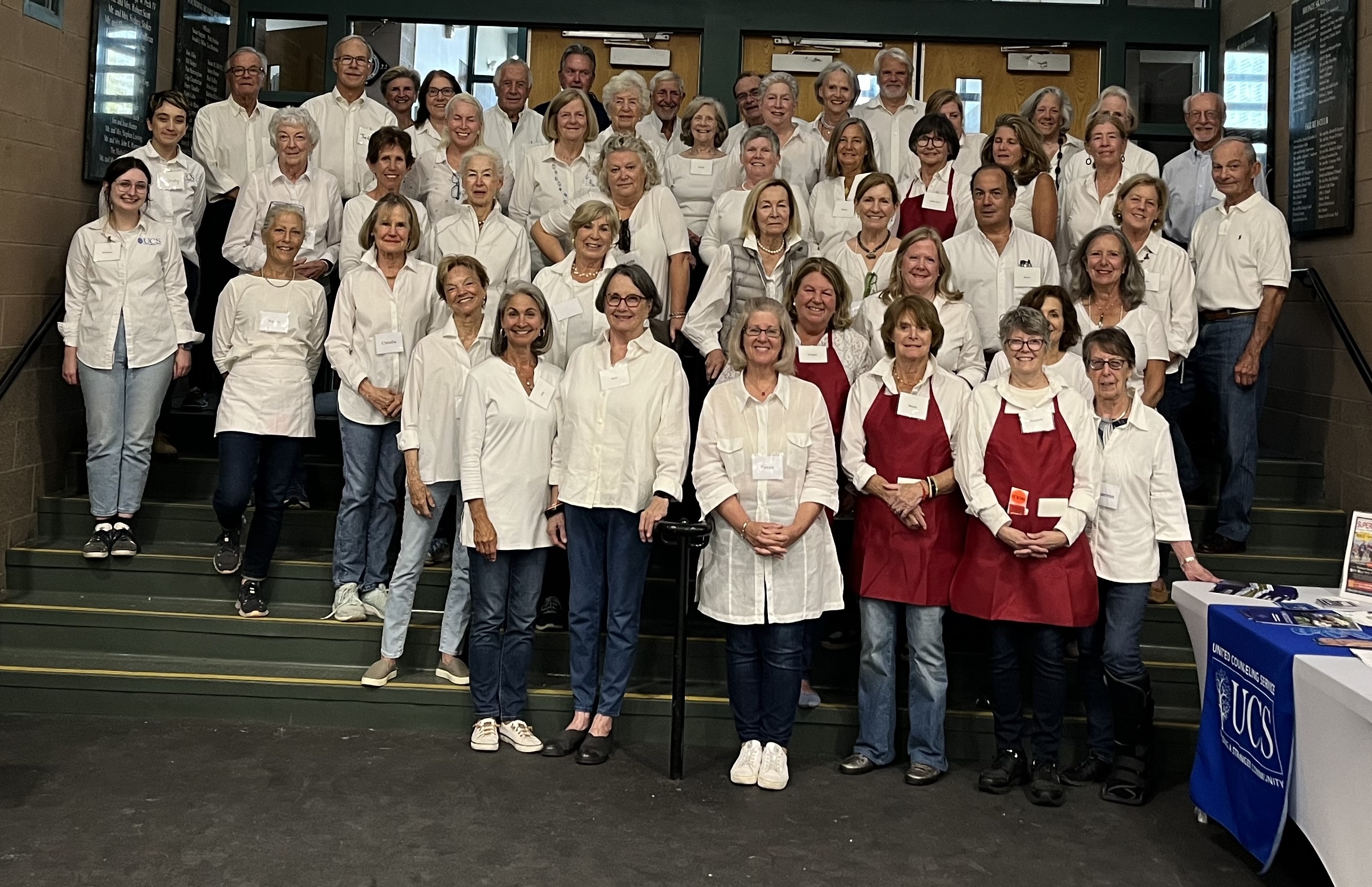 A large group of women and men in white shirts and blue pants stand on steps. Some are wearing red aprons.