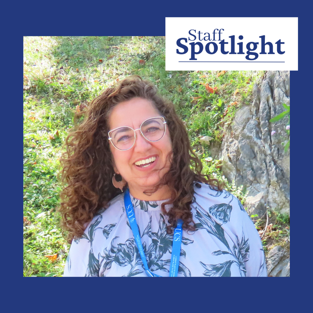 smiling woman with curly brown hair and glasses wearing g=large gold earrings and a light blue dress with darker blue flowers standing against a sunny grassy background. The image is surrounded by a dark blue border with the words Staff Spotlight at the top right.