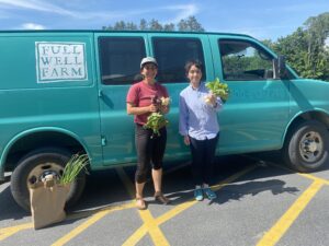 A woman in a red t-shirt,
black capris, and baseball cap and
holding fresh vegetables stands next
to a woman with short black hair, a
blue oxford shirt and black jeans also
holding veggies. They are smiling at
the camera, standing in front of a teal
van with a logo reading “Full Well
Farm”