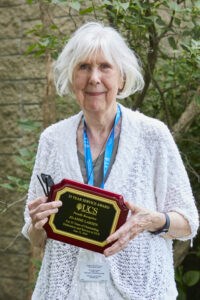 Elder woman with white hair stands outside posing with a plaque