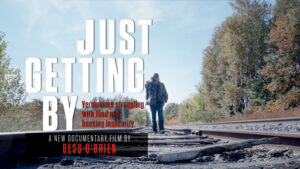 A promo
photo for the documentary Just
Getting By showing a solitary man
walking along abandoned railroad
tracks flanked by fall foliage and a
clear blue sky