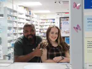 A black man giving a
thumbs-up and a white woman, both
smiling, are leaning on the service
counter looking out from a room of
pharmacy shelves filled with bottles
and boxes