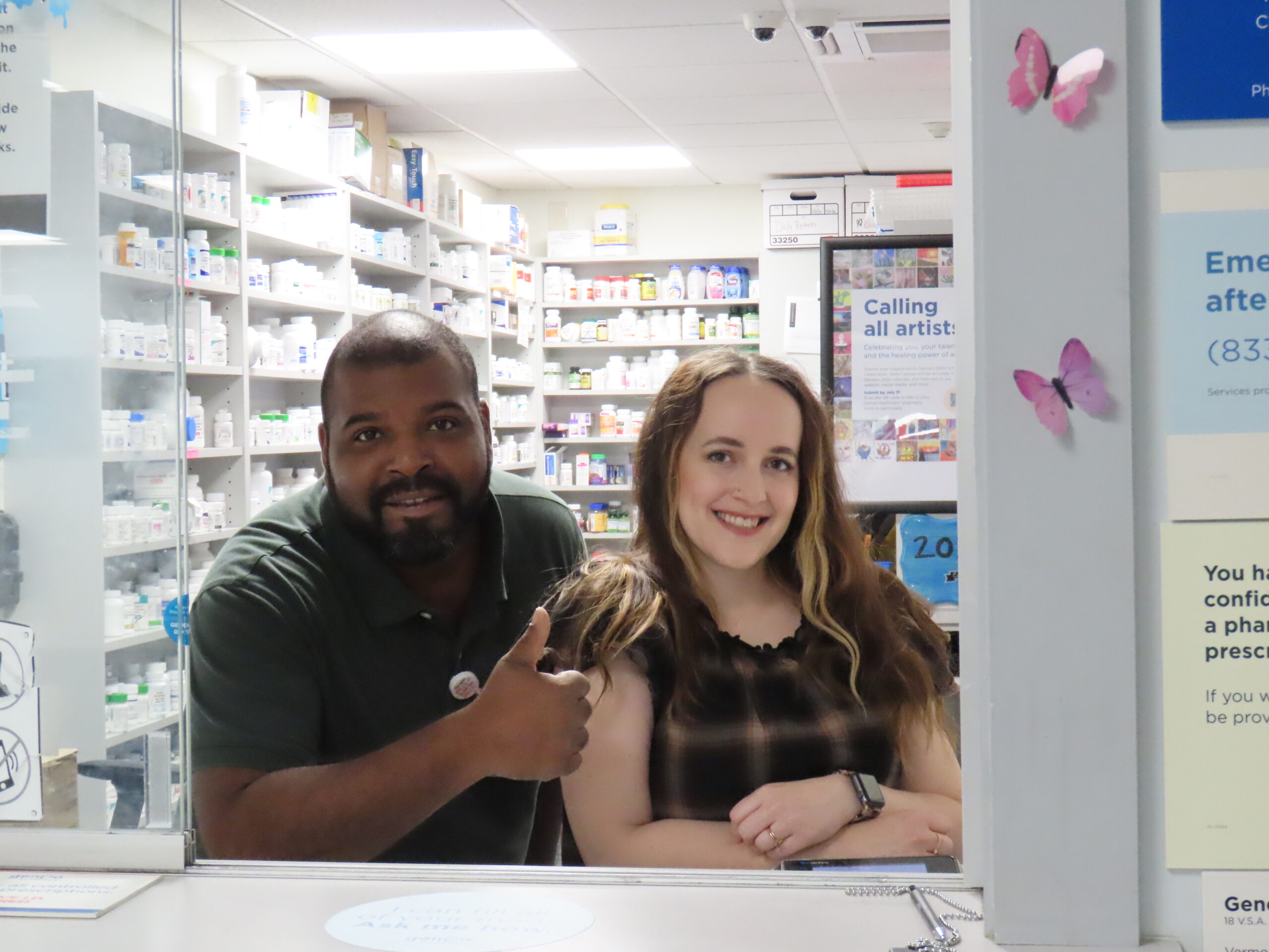 A black man giving a thumbs-up and a white woman, both smiling, are leaning on the service counter looking out from a room of pharmacy shelves filled with bottles and boxes