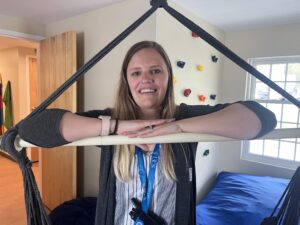 a young woman with long blonde hair and a navy cardigan over a white blouse leans on the bar of a ceiling-mounted trapeze swing. On the wall behind her are colorful plastic pieces mounted to a wall for indoor climbing.