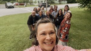 Selfie of a broadly smiling
brown-haired woman in front of a
group of about a dozen smiling
women and two smiling men, all
standing on a lawn surrounding a road
sign.