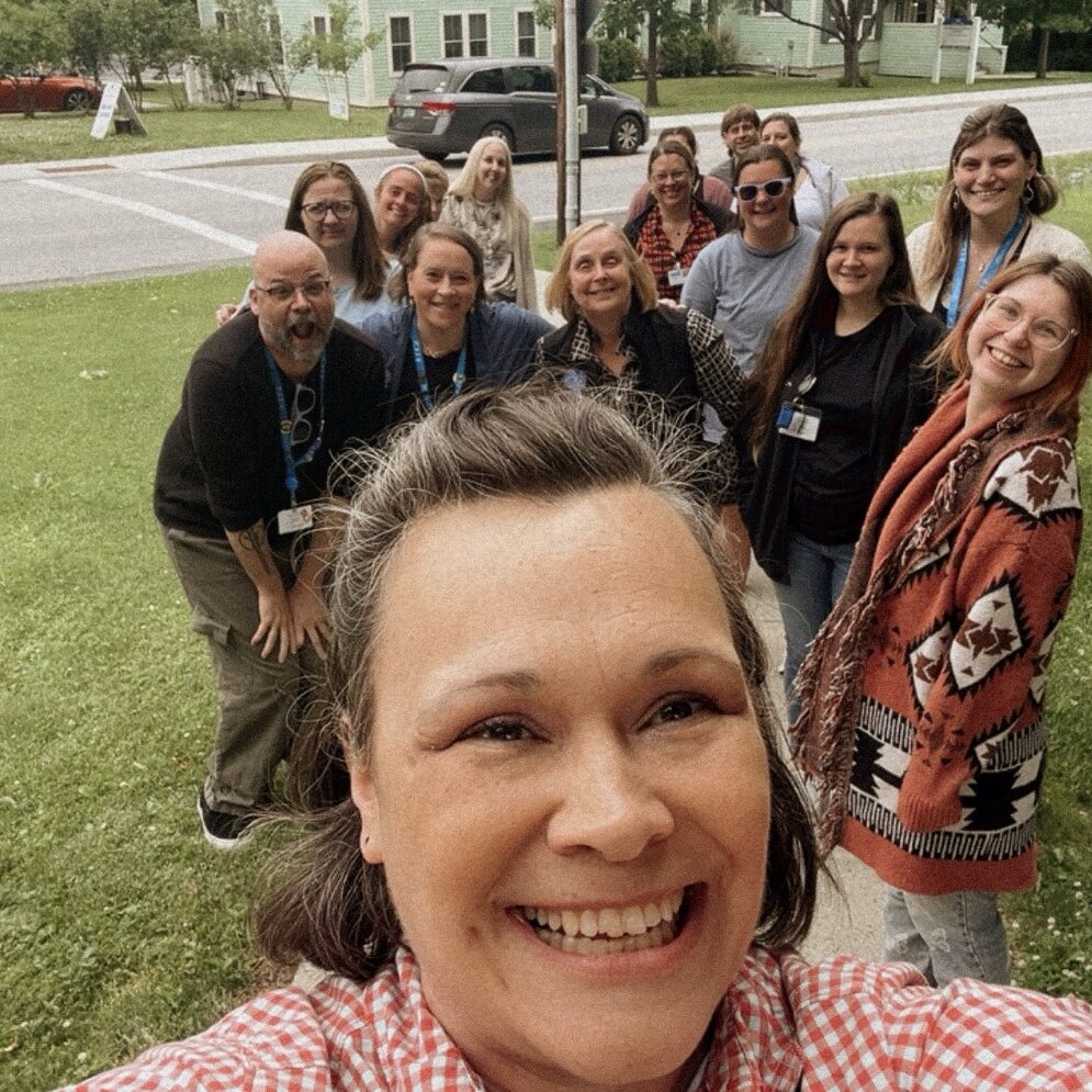 Selfie of a broadly smiling brown-haired woman in front of a group of about a dozen smiling women and two smiling men, all standing on a lawn surrounding a road sign.