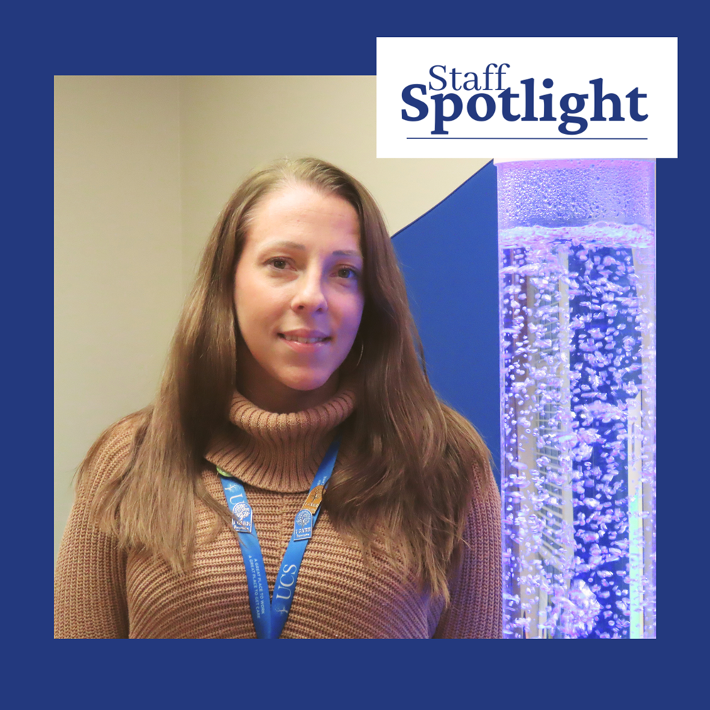 A woman with long brown hair wearing a light brown turtleneck sweater smiles while standing next to a large tube with a liquid and bubbles. There is a dark blue border around the image with a logo overlay that reads Staff Spotlight.