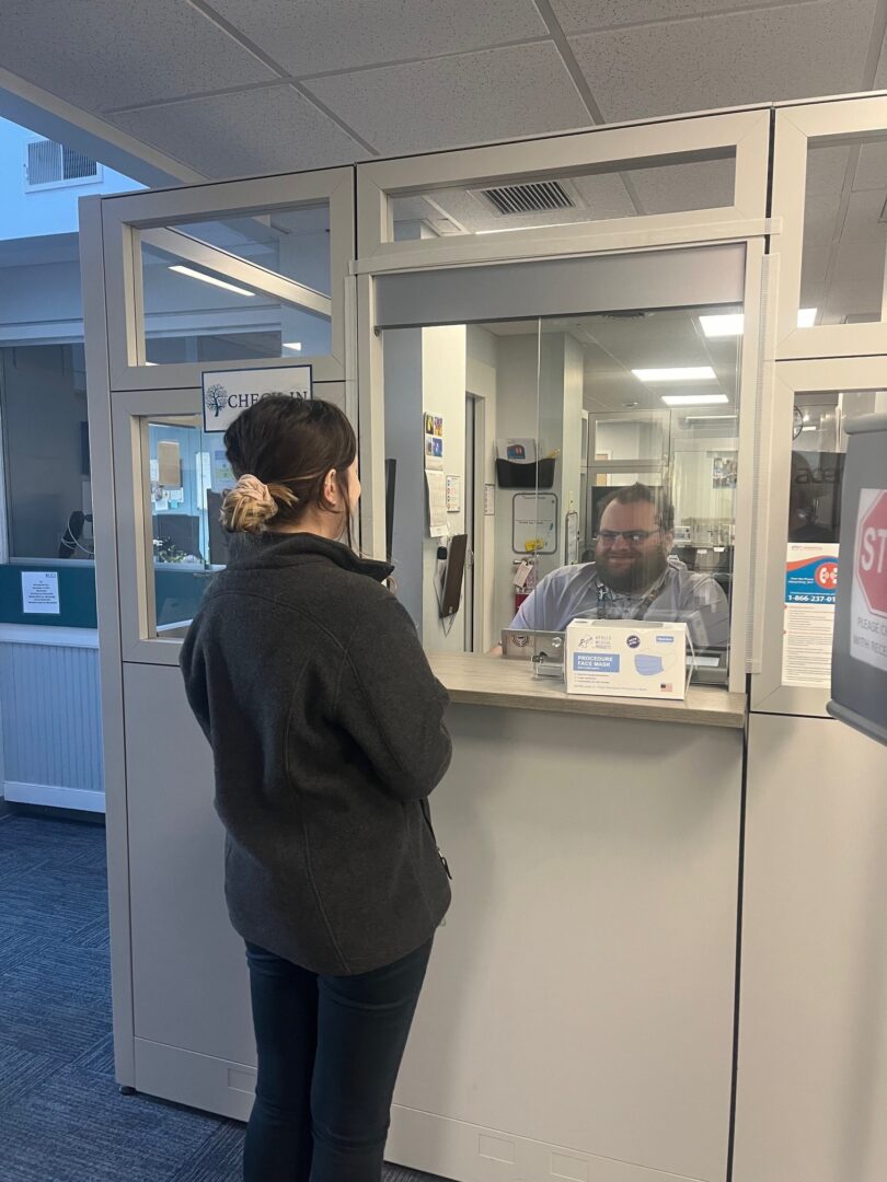 Girl with light brown hair standing at check in desk and greeted by young man with brown hair and a smile.